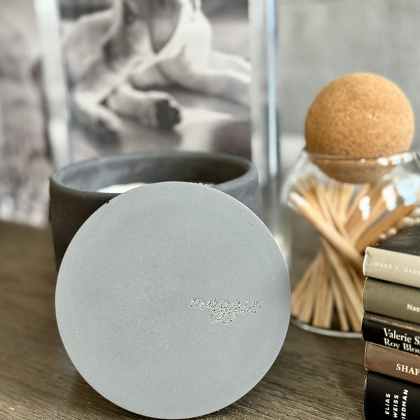 Custom concrete candle in charcoal gray. An example of the engraved lid shows the dog naemorial came, "Magge" in an elegant script font. Each concrete candle is 5.5" in diameter and 3.5" deep with three wicks to burn evenly. Choose a fragrance sucj as Rolling in the grass, Big Stick and also Unscented. Makes the perfect custom pet memorial gift.