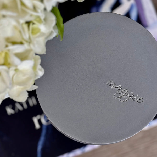 Custom concrete candle in charcoal gray. An example of the engraved lid shows the dog naemorial came, "Magge" in an elegant script font. Each concrete candle is 5.5" in diameter and 3.5" deep with three wicks to burn evenly. Choose a fragrance sucj as Rolling in the grass, Big Stick and also Unscented. Makes the perfect custom pet memorial gift.